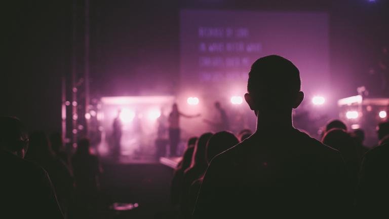 Silhouette of a person in a darkened audience facing a stage with bright purple and white lights. The stage features performers and a large screen with blurry text. The atmosphere suggests a concert or live performance
