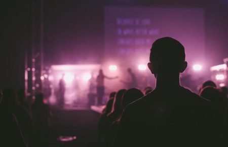 Silhouette of a person in a darkened audience facing a stage with bright purple and white lights. The stage features performers and a large screen with blurry text. The atmosphere suggests a concert or live performance